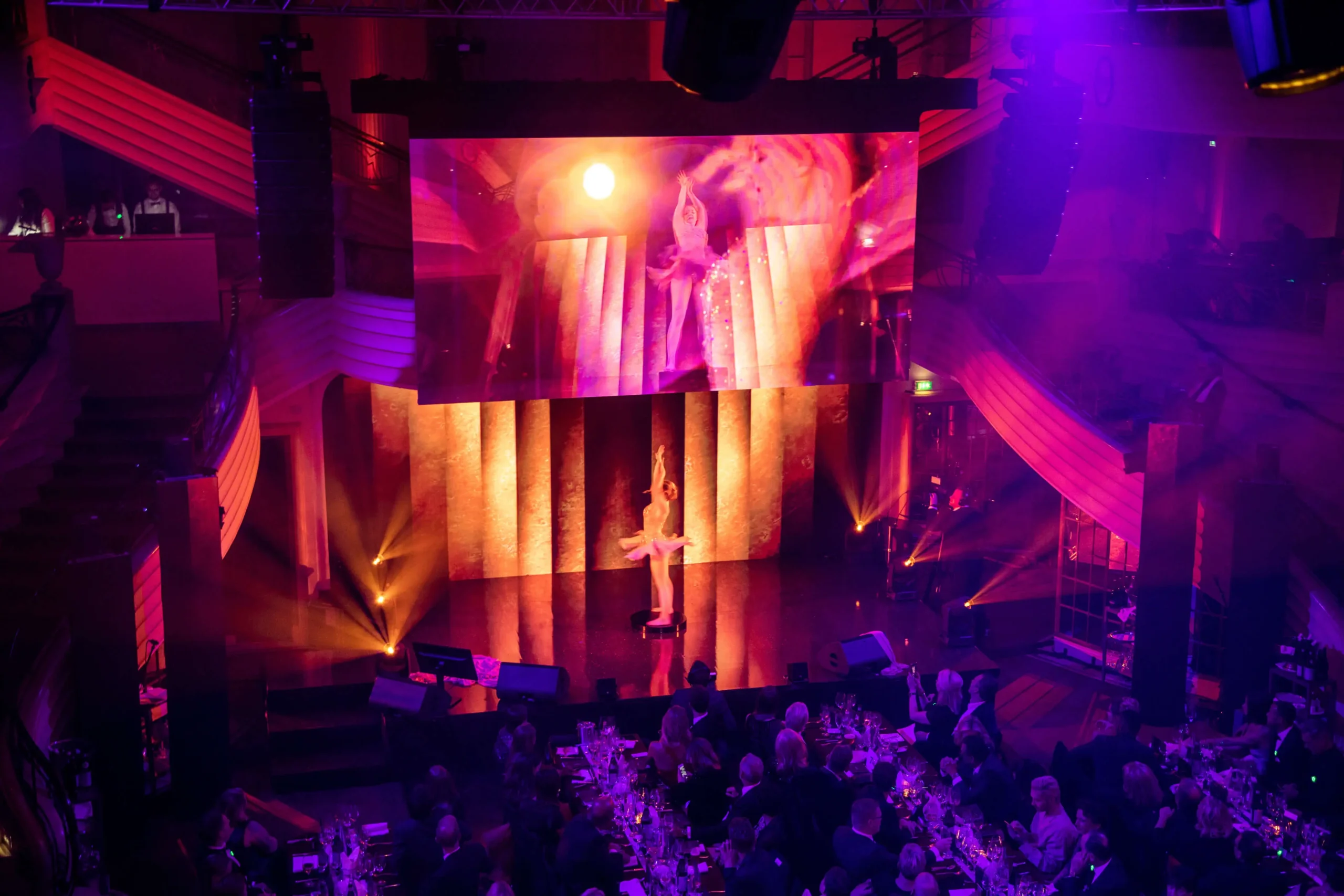 Auf der Bühne der Mc Donalds - CIRCLE SHOW bewegt sich ein Künstler unter orangefarbenen und violetten Lichtern, während eine große Leinwand über ihm sein Bild spiegelt. Die Zuschauer sitzen an Tischen im schwach beleuchteten Vordergrund und lassen sich von dem Spektakel fesseln.