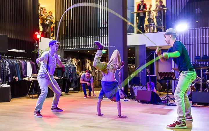 Drei junge Männer zeigen ihr Supertalent, indem sie eine Breakdance-Routine mit doppelten holländischen Springseilen in einer stilvollen Halle vorführen, umgeben von Kleiderständern und Schaulustigen, von denen einige von einem Balkon aus zusehen.