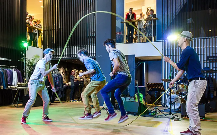 Vier junge Männer zeigen ihr Supertalent im doppelten Seilspringen in einer Halle, wobei zwei die Seile drehen und zwei springen, während ein Publikum sowohl vom Erdgeschoss als auch von einem oberen Balkon aus zusieht. Im Hintergrund sind Musikinstrumente zu sehen.