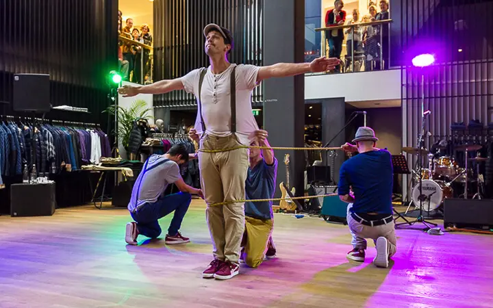 Vier Männer treten in einem Innenraum in einer Supertalent-Nummer auf; einer steht mit ausgestreckten Armen, während drei knien und ein gelbes Seil um seine Taille schlingen. Im Hintergrund sind Einkäufer und Musikinstrumente zu sehen.