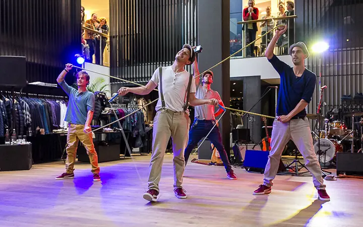 Vier Männer führen einen koordinierten Supertalent-Tanz mit Seilrequisiten in einem modernen Innenraum auf, umgeben von Kleiderständern und Musikinstrumenten, während das Publikum von einem Balkon aus zusieht.