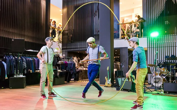 Drei Personen führen in einer Halle beeindruckende Seilsprungtricks vor und zeigen damit ihr Supertalent. Zwei halten ein großes Seil, während der Dritte in der Mitte springt. Lässige Kleidung und Hüte vervollständigen ihren Look, während die Zuschauer von oben zuschauen, mit Kleiderständern und Instrumenten hinter ihnen.