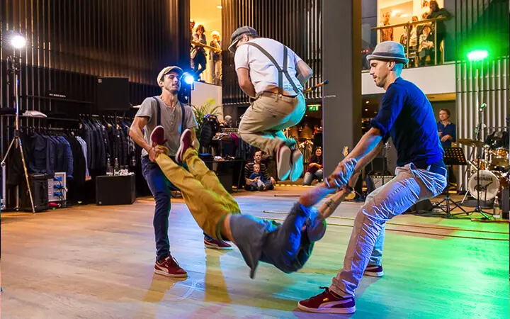 Vier Männer führen einen energiegeladenen Supertalent-Tanz in der Halle auf, wobei einer in der Luft schwebt und ein anderer an Armen und Beinen geschwungen wird. Das Publikum schaut von oben und von hinten zu, während bunte Lichter die lebhafte Szene erhellen.