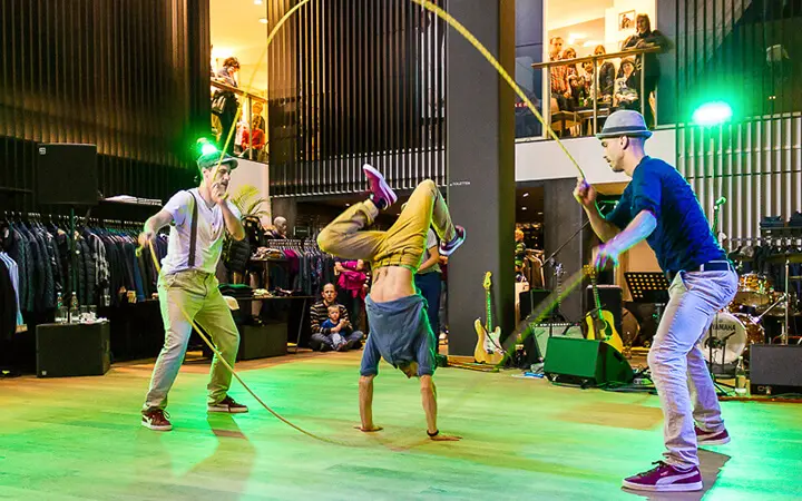 Drei Personen führen einen Supertalent-Seilsprungtrick in einer Halle vor; zwei drehen das Seil, während die dritte einen Handstand in der Mitte macht. Im Hintergrund sind unter bunten Lichtern Musikinstrumente und Schaulustige zu sehen.