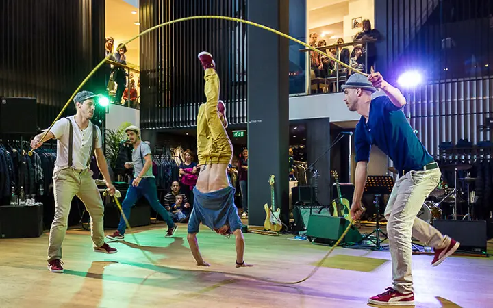 Drei Männer führen eine Supertalent-Seilsprungnummer in einer Halle auf; zwei schwingen das Seil, während der dritte einen Handstand in der Mitte macht. Im Hintergrund sind Zuschauer und Musikinstrumente zu sehen.