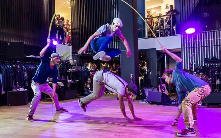 Vier Männer führen ein Double Dutch Jump Roping in der Halle durch; zwei drehen die Seile, während einer hoch springt und ein anderer in die Hocke geht und sein Supertalent in einer dynamischen, energiegeladenen Szene unter Beweis stellt. Die Zuschauer schauen von einem Balkon im Hintergrund zu.