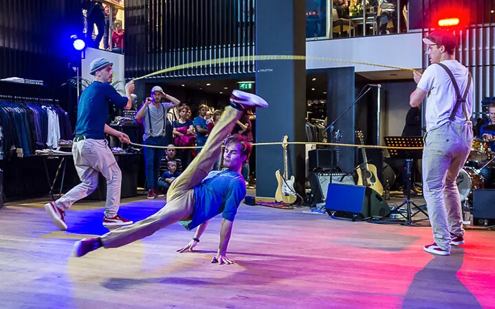 Ein Breakdancer führt eine dynamische Bewegung auf dem Boden aus, während zwei andere auf der Supertalent-Bühne daneben stehen und zusehen, während im Hintergrund Musikinstrumente und eine Zuschauermenge zu sehen sind.