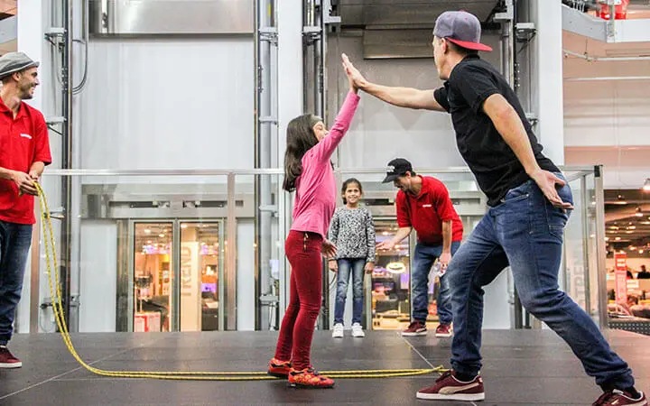 Ein Mann mit einer grauen Mütze gibt einem Mädchen in Rosa auf der Bühne ein High-Five, während zwei andere Männer und ein Mädchen hinter ihnen stehen. Ein Mann, wahrscheinlich ein Rope Skipper, hält ein gelbes Springseil. Die Szene scheint in einem Innenraum zu spielen, möglicherweise bei einer Veranstaltung.