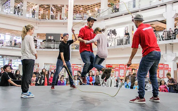 Vier Seilspringer führen auf einer Bühne in einem Einkaufszentrum einen doppelten holländischen Seilsprung durch, wobei eine Menge Zuschauer von mehreren Ebenen aus zusieht. Zwei Personen drehen die Seile, während zwei Seilspringer in der Mitte springen.