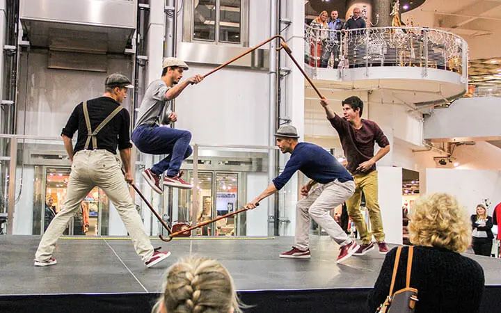 Vier Männer, alle mit Hüten und roten Turnschuhen, führen auf einer Hallenbühne eine koordinierte Ropeskipper-Seilsprungnummer auf. Zwei drehen lange Seile, einer springt gekonnt, während ein anderer darauf wartet, mitzumachen, während das Publikum gespannt zusieht.