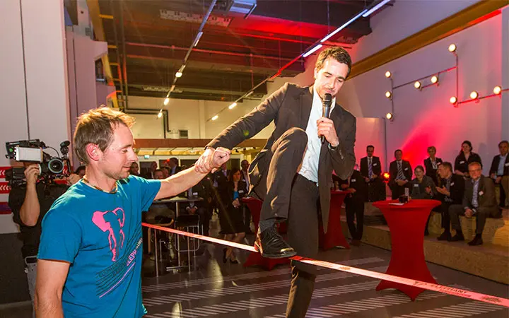 Ein Mann im Anzug, der ein Mikrofon in der Hand hält, versucht während einer mitreißenden Slackline-Show auf einer Slackline zu balancieren. Er wird dabei von einem anderen Mann unterstützt, während das Publikum in einem hell erleuchteten Veranstaltungsraum mit roter Akzentbeleuchtung zusieht.