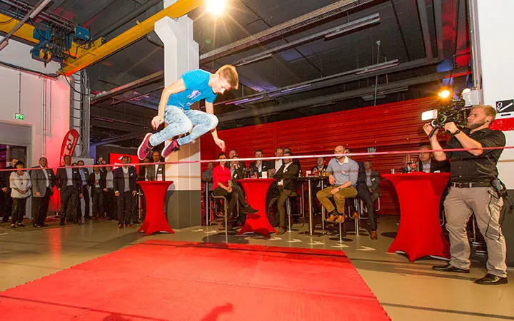 Ein junger Mann in einem blauen Hemd springt während einer Slackline-Show über eine horizontale Stange, während eine Gruppe von Zuschauern und ein Kameramann die Szene filmen. Rote Tische und helle Beleuchtung schmücken den Raum.