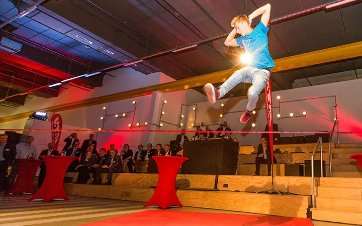 Ein junger Mann in einem blauen Hemd springt während einer aufregenden Slackline-Show in einer Halle über eine hohe Reckstange, während das Publikum auf Holzbänken und rot beleuchteten Tischen im Hintergrund Platz nimmt.