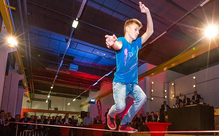 Ein junger Mensch in blauem Hemd und Jeans balanciert mit ausgestreckten Armen auf einer Slackline in einer Halle, während das Publikum im Hintergrund unter bunten Lichtern zusieht.