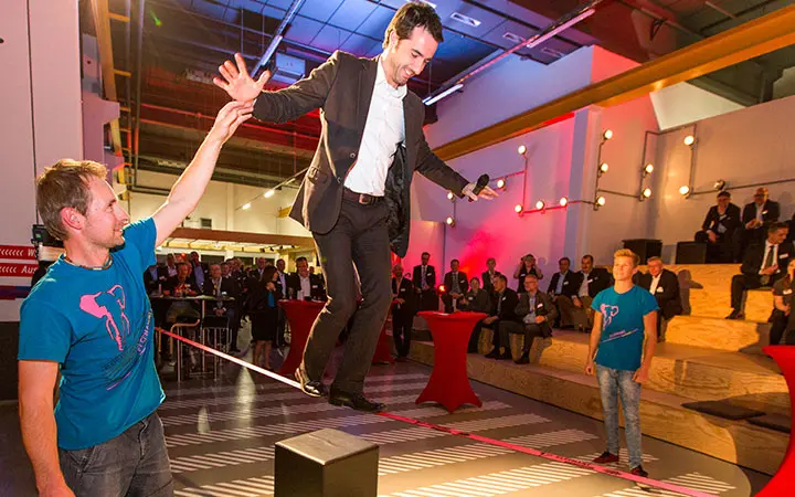 In einem hell erleuchteten Veranstaltungsraum führt ein Mann im Anzug eine Slackline-Show vor. Er balanciert mit einem Mikrofon auf dem Seil, während er sich von einem Mann im blauen Hemd helfen lässt, während die Leute im Hintergrund zusehen.