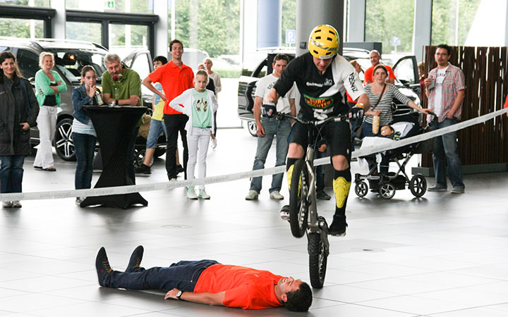 Ein Radfahrer mit Helm vollführt einen waghalsigen Trial-Bike-Sprung über eine am Boden liegende Person in einer Halle, während eine Menschenmenge zusieht und einige Fotos machen.