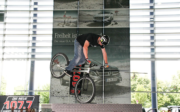 Eine Person, die einen Helm trägt, vollführt einen Balanceakt auf einem Trial-Bike in einer Halle, mit einer Hand am Lenker und einem Fuß auf den Pedalen. Im Hintergrund sind ein Mercedes-Benz Plakat und helle Fenster zu sehen.