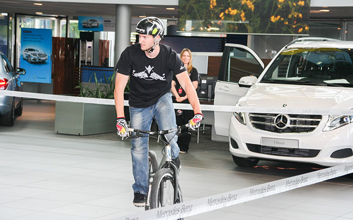 Ein Mann mit Helm und Handschuhen fährt in einer Halle auf einem Trial-Bike, neben ausgestellten weißen Mercedes-Benz Autos. Eine Frau steht im Hintergrund und lächelt. Der Schriftzug Mercedes-Benz erscheint auf einem Band im Vordergrund.