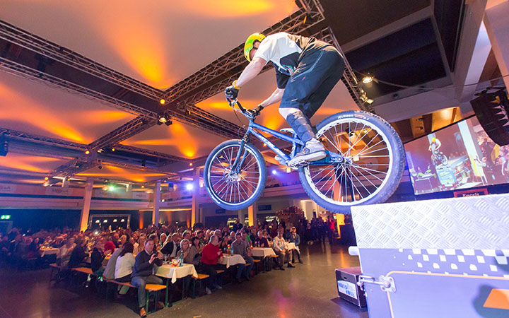 Bei einer Fahrradmesse vollführt ein behelmter Radfahrer auf der Bühne einen waghalsigen Stunt vor einem Publikum, das an Tischen sitzt, die von einer leuchtenden orangefarbenen und blauen Beleuchtung im Veranstaltungsraum angestrahlt werden.