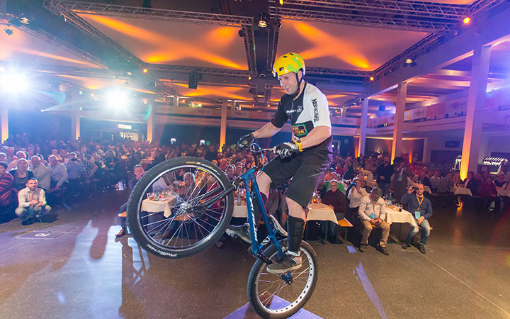 Ein Radfahrer mit gelbem Helm vollführt auf der Fahrradmesse einen Stunt auf einem Mountainbike und balanciert auf einem Rad vor einem sitzenden Publikum unter buntem Bühnenlicht.