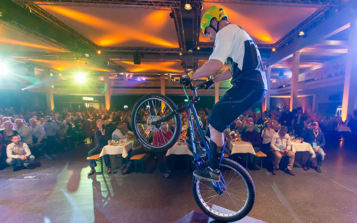 Bei einer Fahrradmesse vollführt ein Radfahrer mit Helm auf einer Bühne im Innenraum einen aufregenden Stunt auf einem Mountainbike, während ein großes, sitzendes Publikum bei bunter Beleuchtung zusieht.