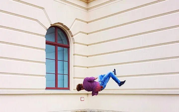 Eine Person in einer lila Jacke und blauen Jeans scheint horizontal in der Luft zu schweben und zeigt ihre Laufkünste vor der beigen Außenwand eines Gebäudes mit einem großen Bogenfenster.