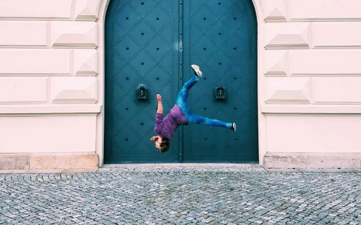 Eine Person in lila Hemd und blauer Jeans führt einen Salto in der Luft aus und zeigt dabei ihre Free-Running-Fähigkeiten vor einer großen, blauen, verzierten Tür in einem hellen Gebäude mit Kopfsteinpflaster.