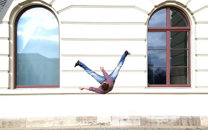 Eine Person, die Free Running praktiziert, scheint seitlich auf einer weißen Gebäudewand zwischen zwei großen Bogenfenstern zu liegen, wobei ihre Beine in der Luft gespreizt sind, was eine optische Täuschung erzeugt, als ob die Schwerkraft seitwärts wirkt.