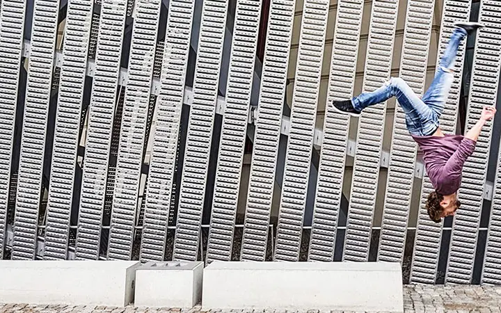 Eine Person, die Jeans und ein langärmeliges Hemd trägt, wird mitten im Rückwärtssalto beim freien Laufen vor einer großen Wand mit gleichmäßig verteilten Metalllatten eingefangen, wodurch eine dynamische und energiegeladene Szene entsteht.