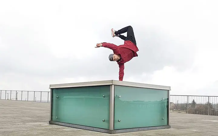 Eine Person in roter Jacke und Mütze zeigt beeindruckende Free-Running-Fähigkeiten, indem sie einen einhändigen Handstand auf einer Glasstruktur in einem offenen Außenbereich unter einem bewölkten Himmel ausführt.