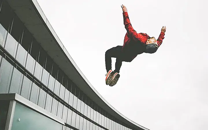 Eine Person in roter Jacke und Turnschuhen zeigt beeindruckende Free-Running-Künste und vollführt einen Rückwärtssalto in der Luft neben einem modernen, geschwungenen Gebäude vor einem bewölkten Himmel.