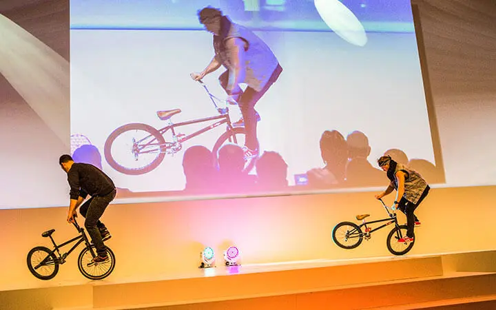 Zwei Personen führen auf einer Bühne BMX-Tricks vor, wobei jeder auf dem Hinterrad eines Fahrrads balanciert. Hinter ihnen wird auf einer großen Leinwand das Bild eines Fahrers projiziert, und die Silhouetten des Publikums sind zu sehen - perfekt für ein Künstlerverzeichnis-Showcase.