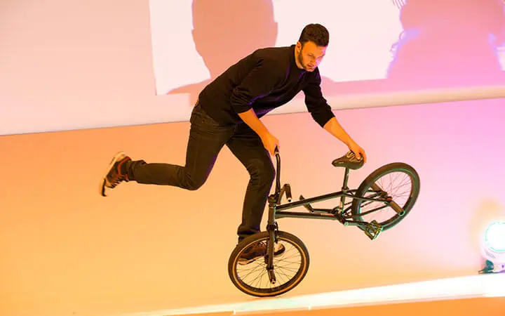 Ein Mann in schwarzem Pullover und Jeans führt einen Trick auf einem BMX-Rad vor, wobei er auf einem Rad balanciert, während ein Fuß vom Pedal genommen wird - perfekt für ein Künstlerverzeichnis, das urbane Künstler zeigt.