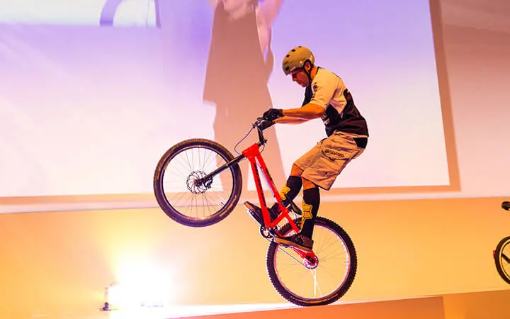 Ein Radfahrer, der in einem Künstlerverzeichnis abgebildet ist, trägt einen Helm und eine Sportausrüstung, während er in einer Halle auf einem roten Mountainbike einen Trick vorführt und dabei auf dem Hinterrad balanciert, während im Hintergrund ein großer Bildschirm und helle Lichter zu sehen sind.