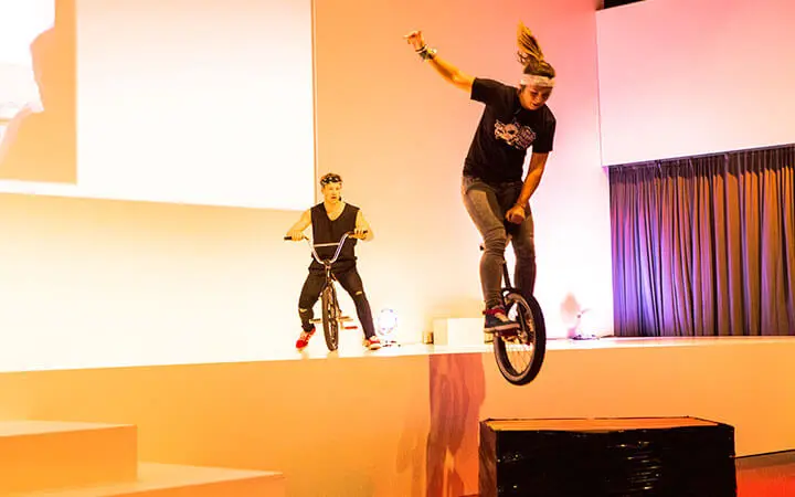 Zwei Personen führen in einer Halle Kunststücke auf Einrädern vor; die vordere Person springt in der Luft von einer Plattform, während die andere im Hintergrund fährt. Die hell erleuchtete Szene mit weißer und rosa Beleuchtung zeigt ein Talent, das eines Künstlerverzeichnisses würdig ist.