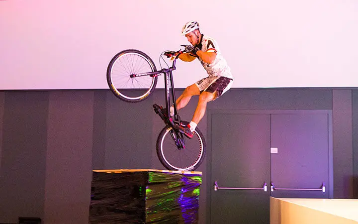 Ein im Künstlerverzeichnis eingetragener Radfahrer, der einen Helm und Sportkleidung trägt, balanciert auf dem Hinterrad eines Fahrrads auf einer erhöhten Plattform in einer Halle, mit Doppeltüren und einer weißen Wand im Hintergrund.