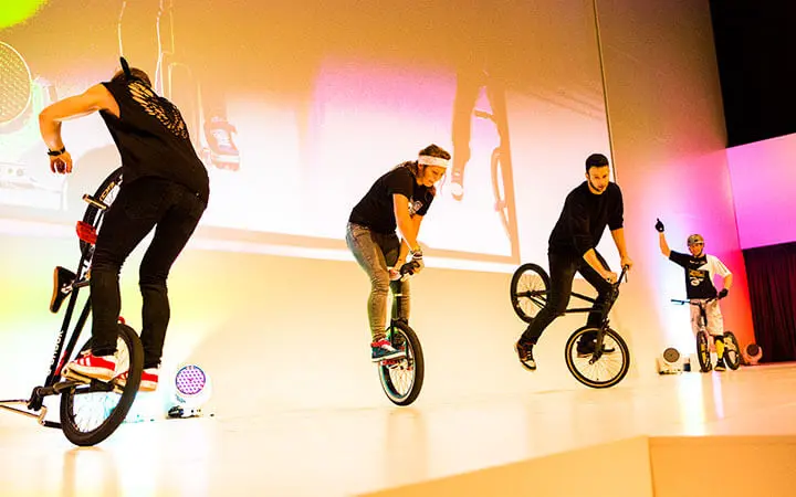 Vier BMX-Fahrer führen auf der Bühne unter bunten Lichtern Tricks vor, wobei drei auf einem Rad fahren und einer im Hintergrund eine Hand hebt. Ihre dynamische Show, die im Künstlerverzeichnis aufgeführt ist, wirft kühne Schatten auf BMX-Grafiken.