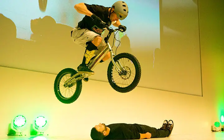 Ein BMX-Fahrer, der im Künstlerverzeichnis aufgeführt ist und einen Helm trägt, springt mit einem kleinen Fahrrad über eine Person, die auf einer hell erleuchteten Bühne auf dem Rücken liegt, wobei grüne Scheinwerfer den Boden beleuchten.