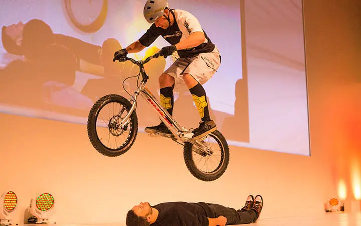 Ein Radfahrer mit Helm, der im Künstlerverzeichnis aufgeführt ist, vollführt einen Stunt, bei dem er mit dem Fahrrad über eine am Boden liegende Person springt, während im Hintergrund helle Lichter und ein projiziertes Bild zu sehen sind.