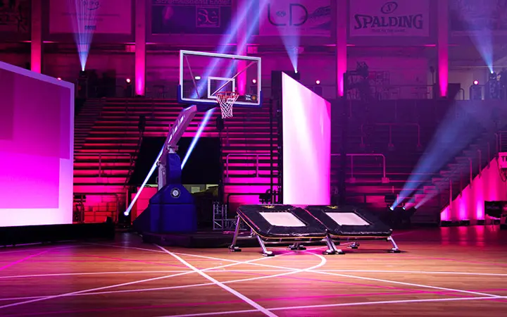 Ein Basketballkorb auf einem Spielfeld wird für eine Slam Dunk Show mit rosa und lila Bühnenscheinwerfern beleuchtet. Vor dem Korb sind zwei Trampoline aufgestellt; im Hintergrund ist eine leere Tribüne zu sehen.