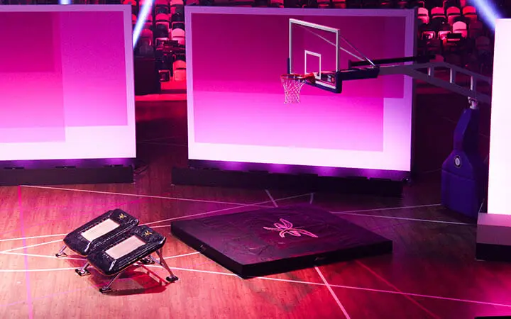 Ein Basketballkorb steht auf einem Spielfeld mit zwei Trampolinen und einer großen Sprungmatte, das für eine aufregende Slam Dunk Show eingerichtet ist, umgeben von rosa und lila Beleuchtung und leeren Zuschauerplätzen im Hintergrund.