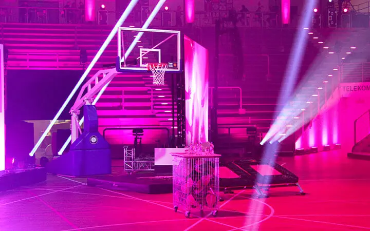 Ein Basketballplatz ist mit hellen rosa und lila Lichtern beleuchtet. In der Mitte steht ein Basketballkorb, um den herum Scheinwerfer und Bühnentechnik aufgebaut sind. So entsteht eine lebendige, ereignisreiche Atmosphäre, die perfekt für eine Slam Dunk Show ist.
