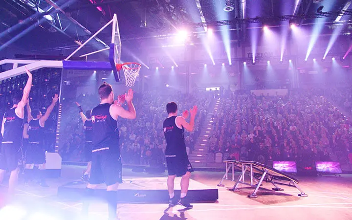 Basketballspieler in schwarzen Trikots treten auf einem hell erleuchteten Spielfeld auf und applaudieren dem zahlreich erschienenen Publikum. Lila Bühnenlichter erhellen die Arena und sorgen für eine lebhafte und energiegeladene Stimmung bei der aufregenden Slam Dunk Show.