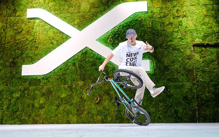 Ein Freestyle-Künstler vollführt einen BMX-Rad-Trick auf einem Rad vor einer grünen Mooswand mit einem großen weißen "X"-Symbol. Der Fahrer trägt ein weißes Hemd, eine beige Hose und eine Kappe.