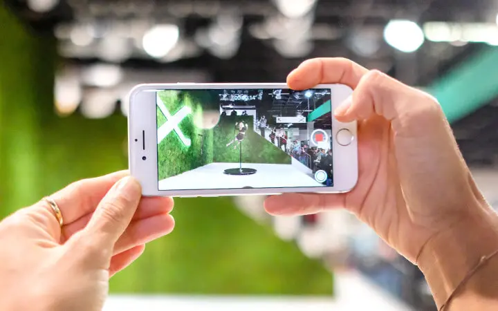 Eine Person hält ein Smartphone in der Hand und nimmt ein Foto oder ein Video von einem Display mit einem grünen Hintergrund und einem großen weißen X auf - vielleicht dokumentiert sie die einzigartige Einrichtung eines Freestyle-Künstlers. Der Bildschirm des Telefons spiegelt die beeindruckende Szene wider.