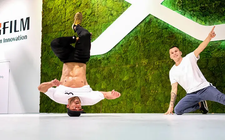 Zwei Männer zeigen in einer Halle ihre Freestyle-Künste; der eine balanciert auf dem Kopf mit den Beinen in der Luft, während der andere neben ihm kniet, lächelt und auf ihn zeigt. Im Hintergrund sind eine grüne Mooswand und das Firmenlogo zu sehen.