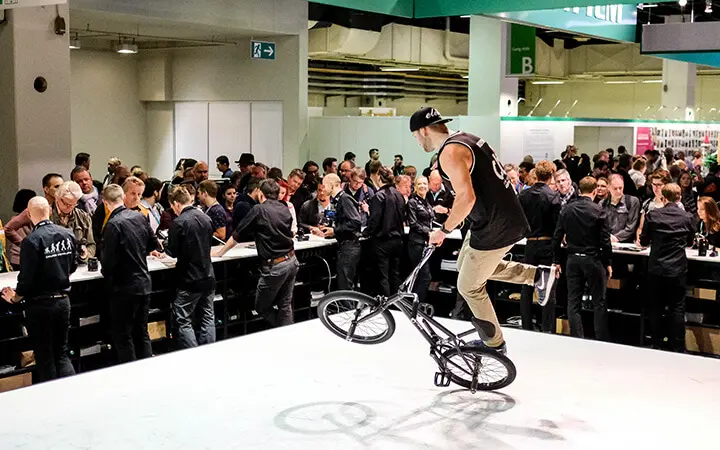 Ein Freestyle-Künstler führt einen BMX-Trick auf der Bühne vor einem großen Publikum vor, das an Tischen in einer Veranstaltungshalle sitzt und steht. Das Publikum schaut aufmerksam zu.