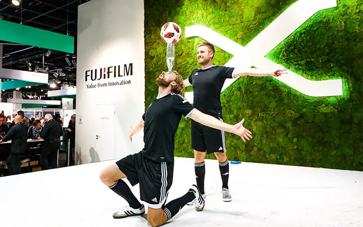 Zwei Freestyle-Künstler in schwarzer Sportkleidung führen einen Fußballtrick vor - einer kniet und balanciert einen Ball auf seinem Kopf, während der andere mit ausgestreckten Armen dahinter steht. Ein FUJIFILM-Schild und eine grüne Wand vervollständigen den Hintergrund.
