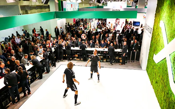 Zwei Freestyle-Künstler zeigen Fußballtricks auf einer weißen Bühne, umgeben von einer Menschenmenge bei einer Indoor-Veranstaltung. Eine große grüne Wand mit einem weißen "X" ist zu sehen, während die Leute von Tischen und der Veranstaltungsfläche aus zusehen.