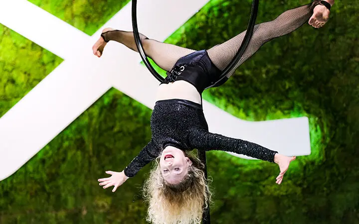 Ein Freestyle-Künstler mit lockigem blondem Haar hängt kopfüber an einem Luftreifen, Arme und Beine ausgestreckt, vor einem grünen Hintergrund mit einem großen weißen X.
